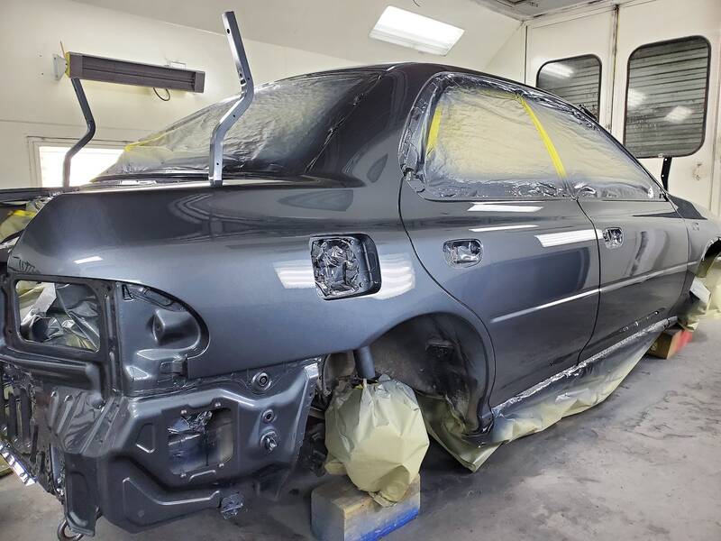 Dark gray sedan in a white paint booth, windows, lights and wheels masked with plastic and yellow tape