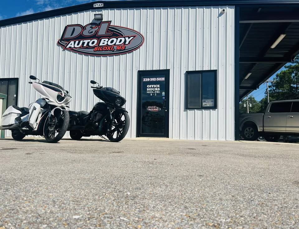 Front of the shop: white metal building with the red-ringed D&L Auto Body oval sign, black office door and two motorcycles parked out front