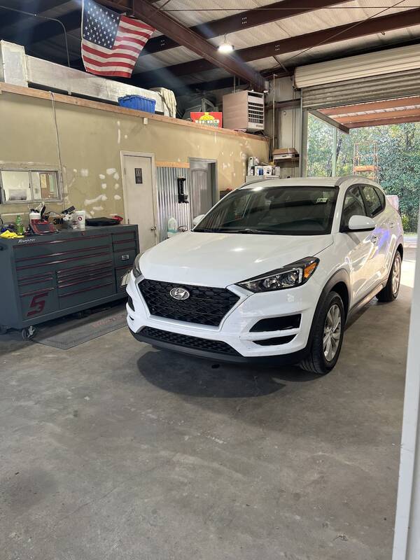 White Hyundai Tucson parked inside the shop bay next to a red-striped tool chest, with an American flag on the wall
